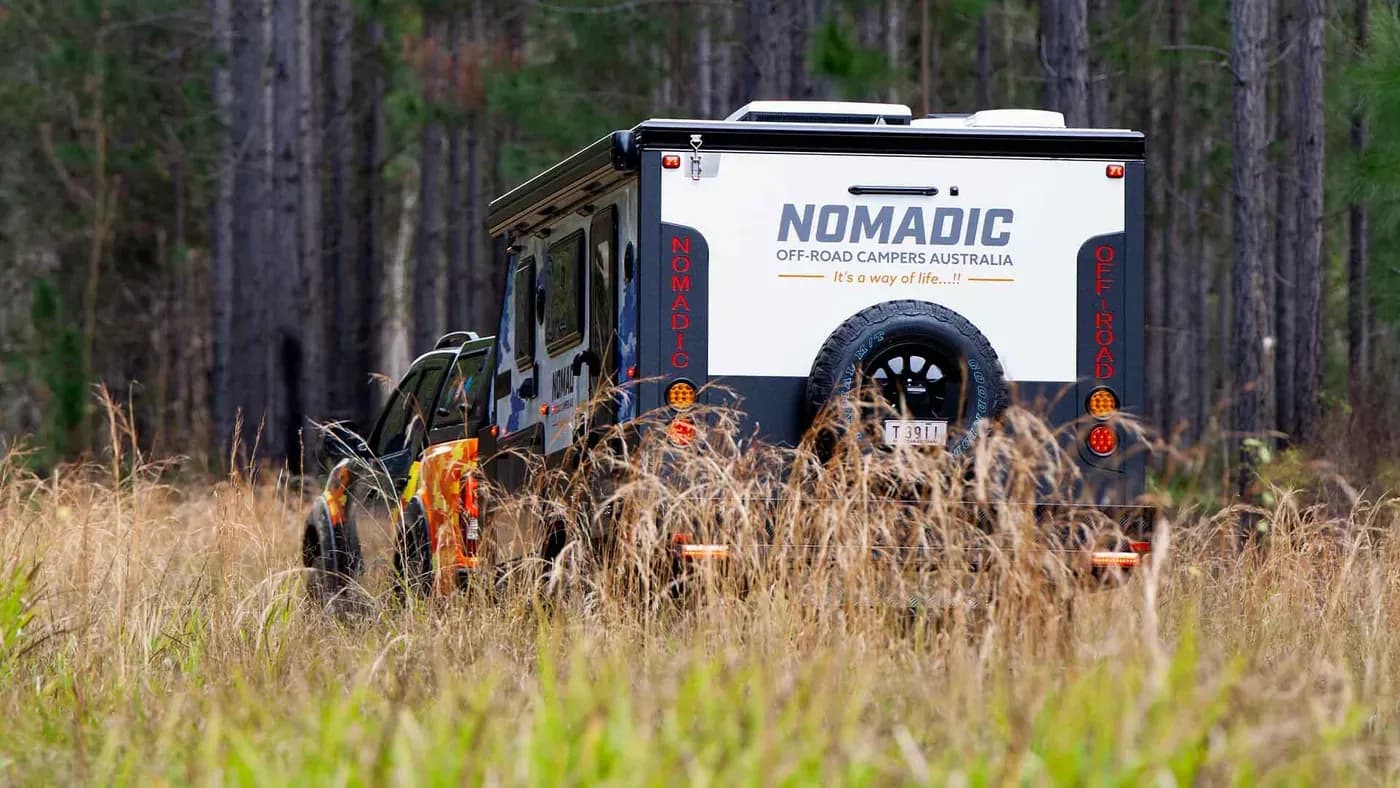 Nomadic off-road camper and tow rig tracking through a pine forest, rear three-quarter view