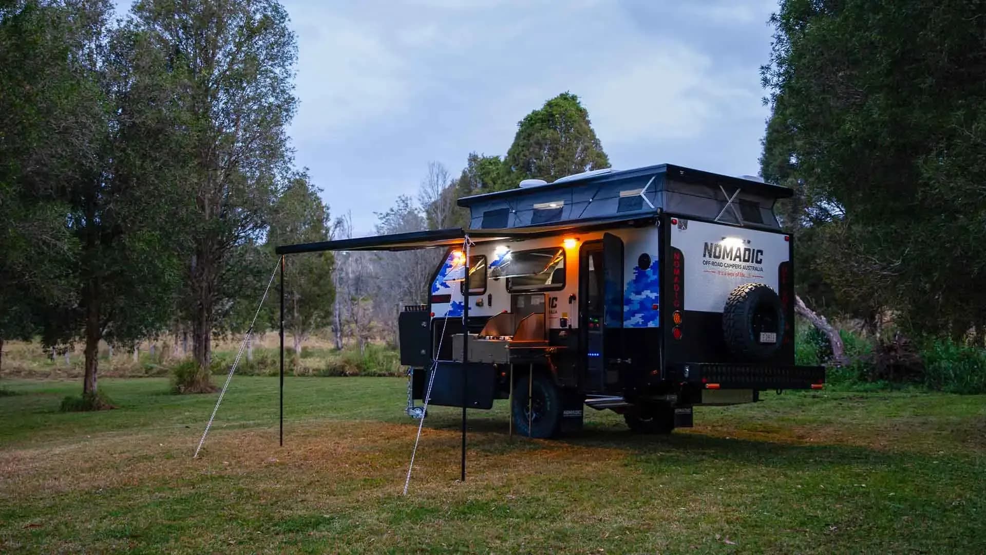Nomadic pop-top camper at a dusk campsite with the awning out and the external kitchen lit
